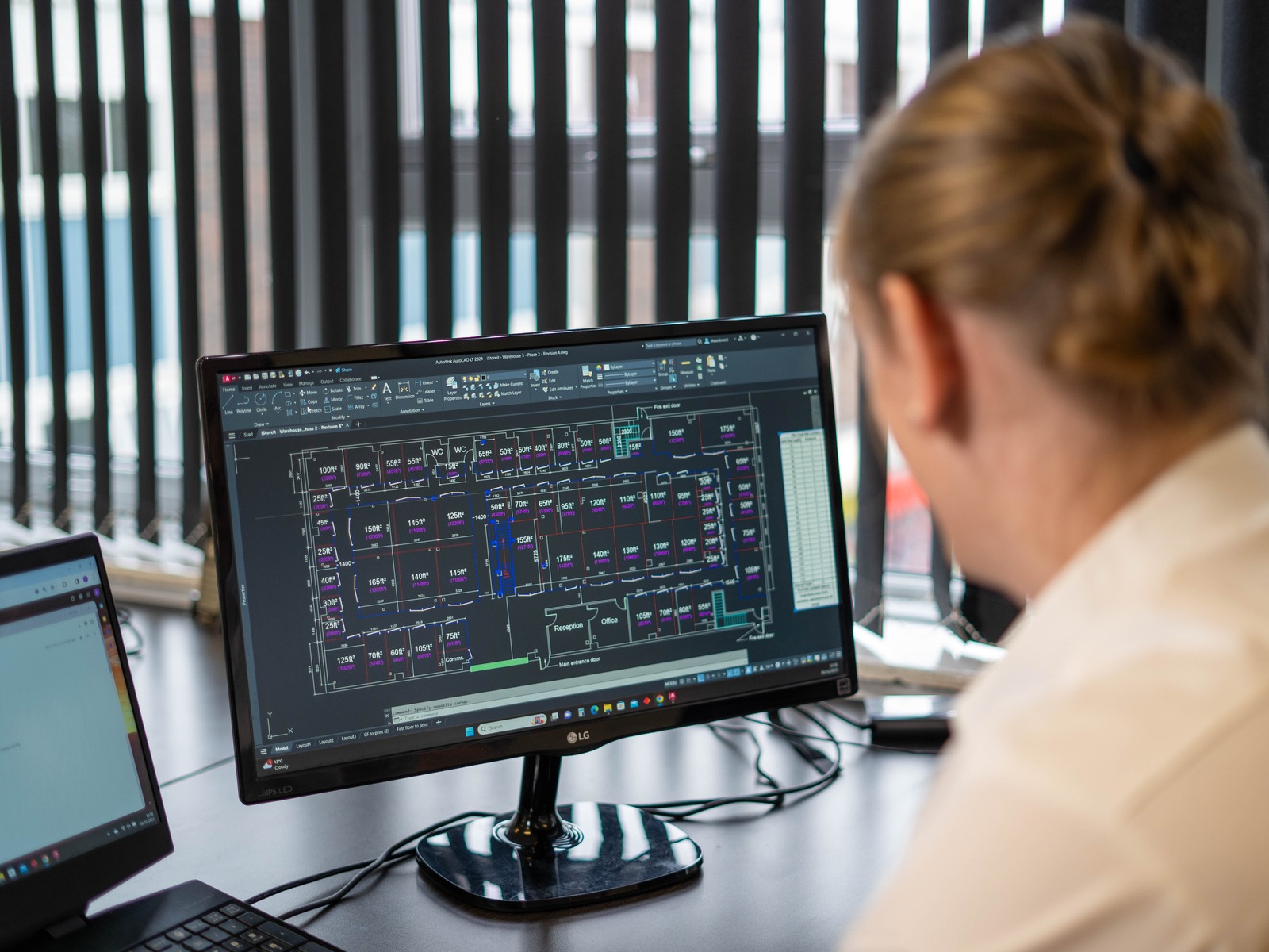 A person with blonde hair in a bun is seated at a desk, looking at a computer monitor displaying a complex schematic or technical drawing. Another laptop is partially visible to the left, and the background has vertical blinds covering windows.