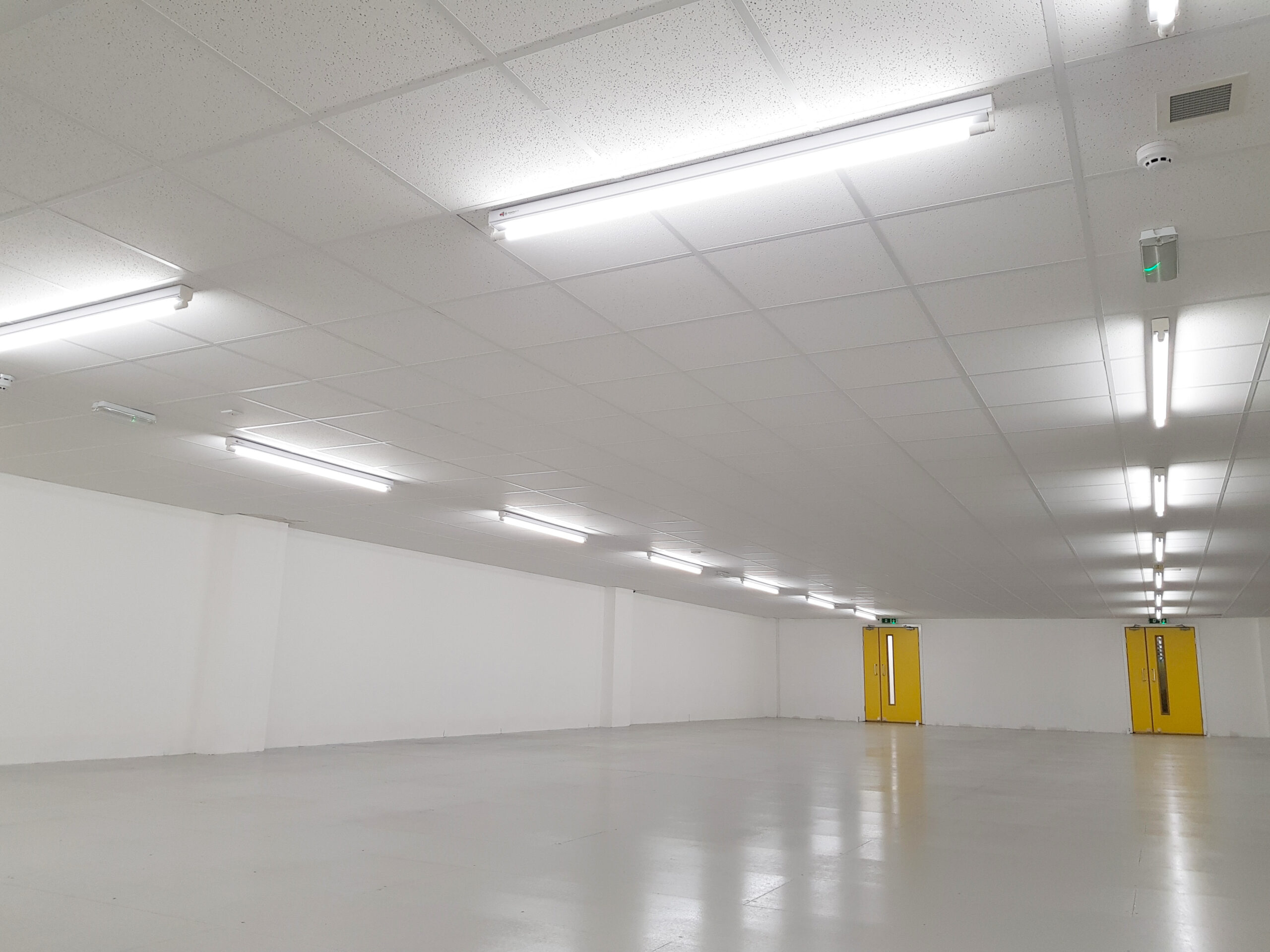 A spacious, empty room with white walls and a white tile ceiling. The floor is clean and smooth, reflecting the overhead fluorescent lights. Two yellow doors are visible on the far wall. The room appears well-lit and has a modern, minimalistic design, ideal for self-storage with integrated fire protection systems.