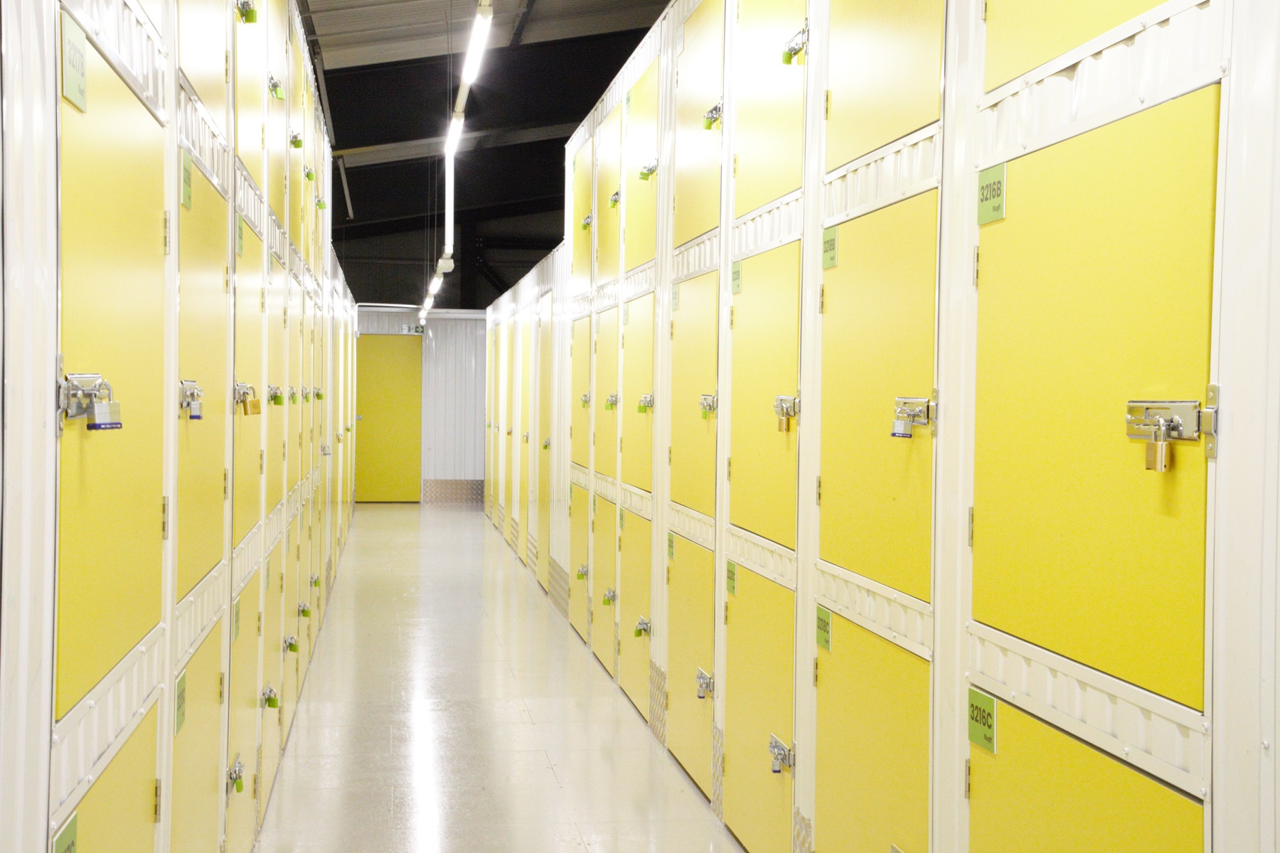 A brightly lit storage facility features yellow locker storage units lining both sides of a long, narrow hallway. Each door is secured with a padlock, and the clean white floor and ceiling panels enhance the orderly appearance of the space.