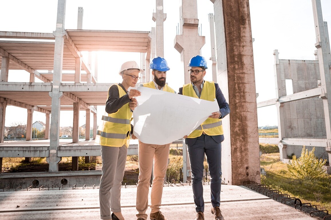 Three construction workers wearing yellow safety vests and blue helmets examine a large blueprint for a storage facility design at a construction site. They are surrounded by concrete structures and beams, with sunlight visible in the background.