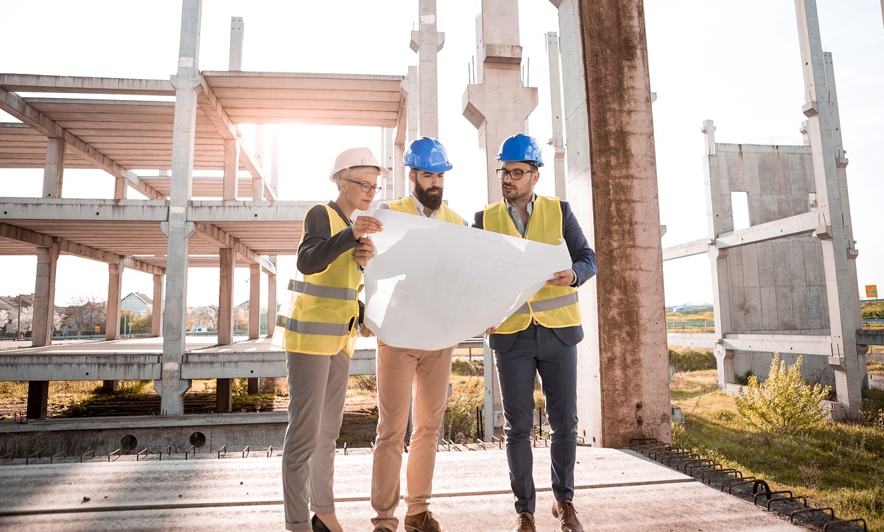 Three construction workers wearing yellow safety vests and blue helmets examine a large blueprint for a storage facility design at a construction site. They are surrounded by concrete structures and beams, with sunlight visible in the background.