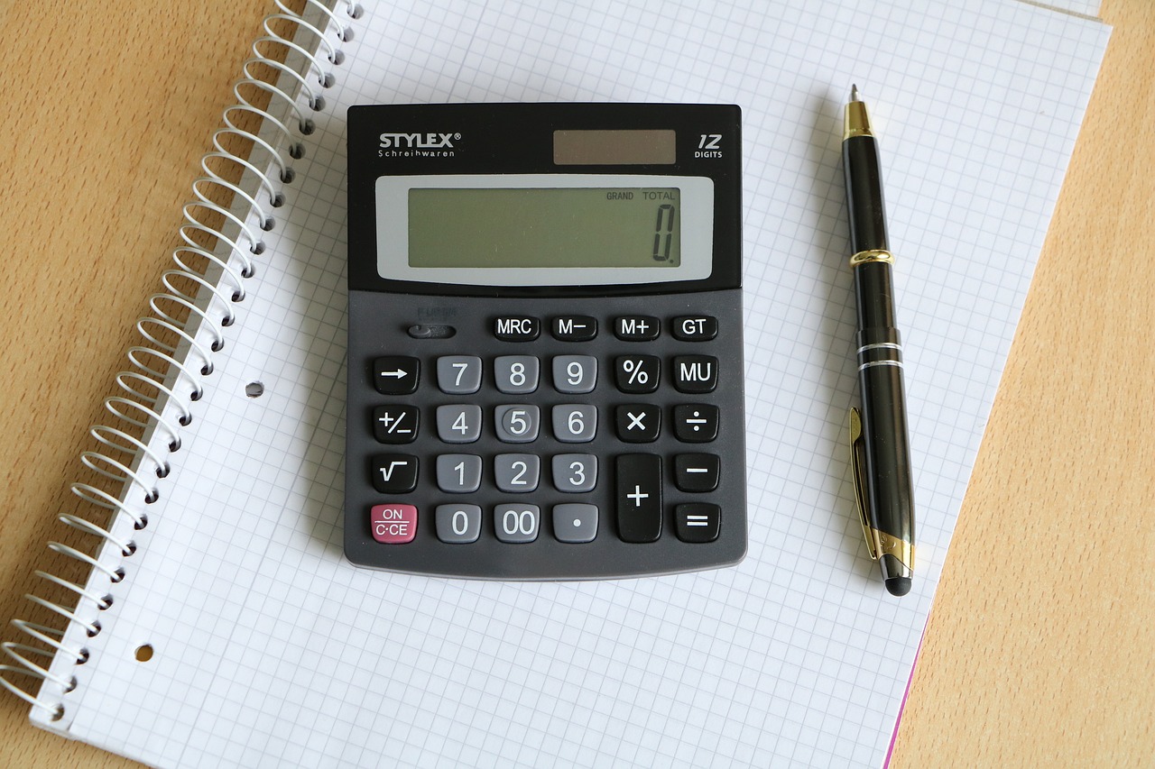 A black and grey calculator lies on top of a graph paper notebook, perfect for tackling financing tasks. A black and gold pen is placed to the right of the calculator. The notebook has a spiral binding along its left edge, positioned on a light-colored wooden surface.