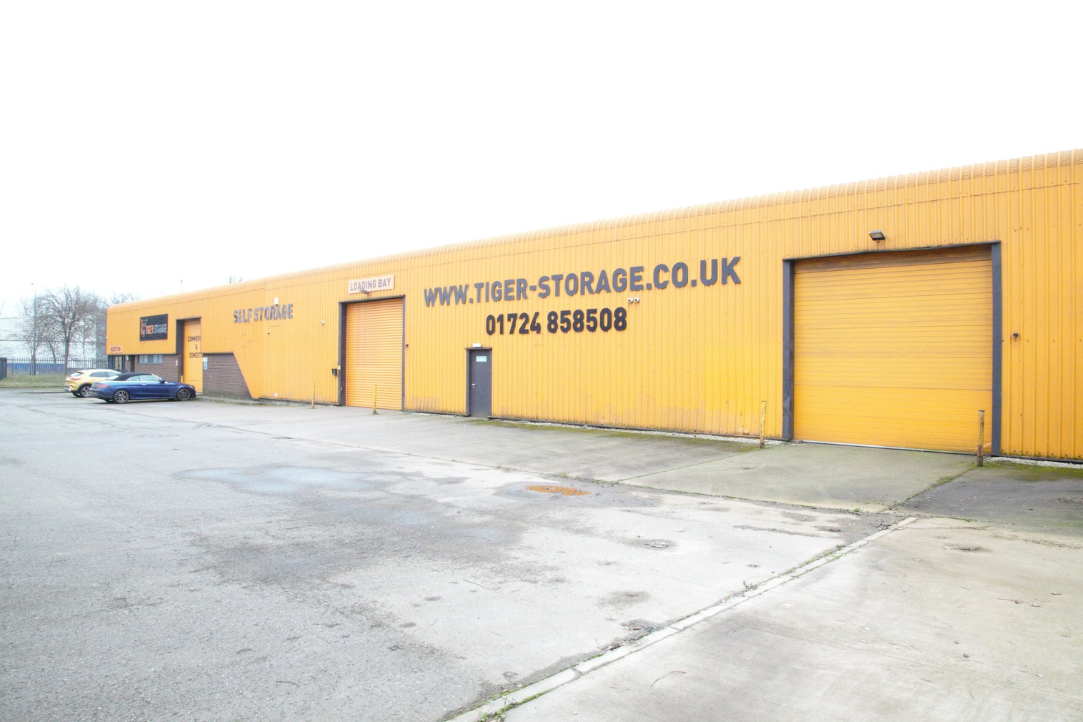 A large yellow industrial building with "www.Tiger-Storage.co.uk" and a phone number written on the side. The facility has several garage doors and a small entrance door. A single blue car is parked near the building. The surrounding area is a paved lot.