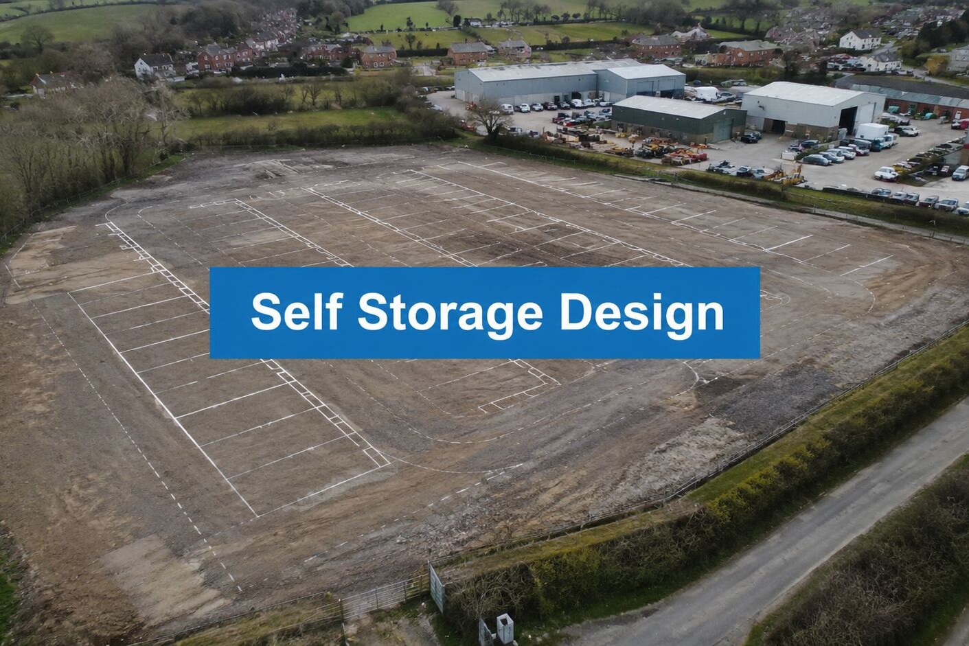 Aerial view of a large, cleared dirt lot with white lines marking sections, bordered by roads and trees, with nearby buildings and parked cars. Text overlay reads “Self Storage Design.”.